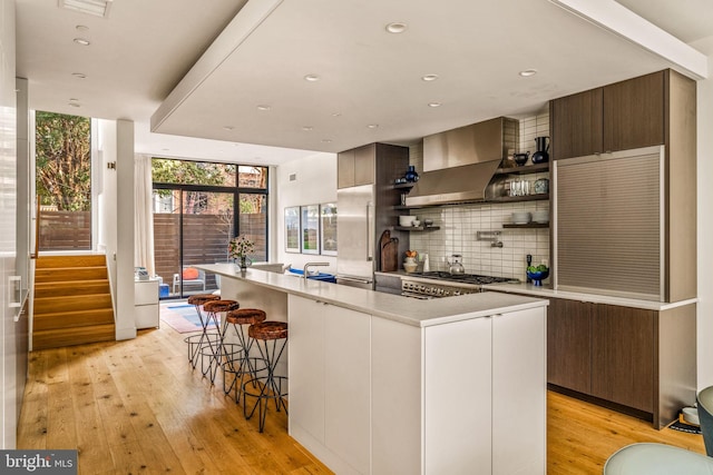 kitchen with open shelves, light countertops, decorative backsplash, a kitchen island with sink, and wall chimney exhaust hood