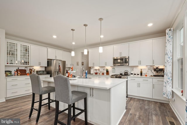 kitchen with a kitchen island with sink, stainless steel appliances, white cabinetry, hanging light fixtures, and glass insert cabinets