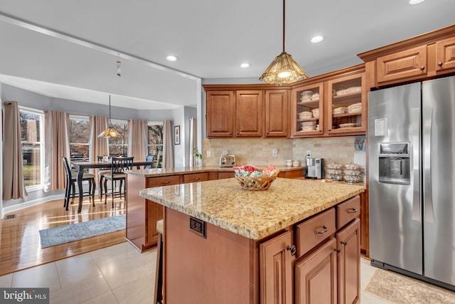 kitchen with light stone countertops, a center island, hanging light fixtures, stainless steel refrigerator with ice dispenser, and kitchen peninsula