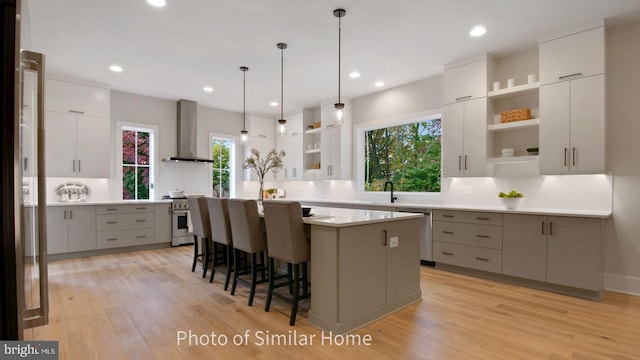 kitchen with pendant lighting, wall chimney range hood, stainless steel appliances, an island with sink, and light hardwood / wood-style floors