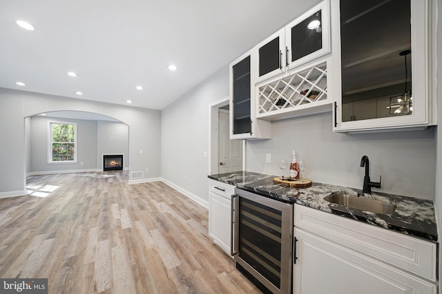 bar with sink, beverage cooler, dark stone countertops, white cabinets, and light wood-type flooring