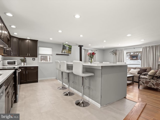 kitchen with stainless steel range with electric cooktop, light wood-type flooring, a breakfast bar area, dark brown cabinetry, and dishwashing machine