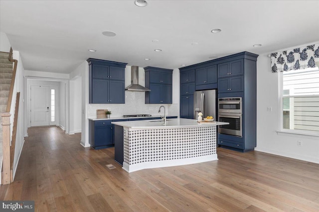 kitchen featuring appliances with stainless steel finishes, light wood-type flooring, an island with sink, and wall chimney range hood