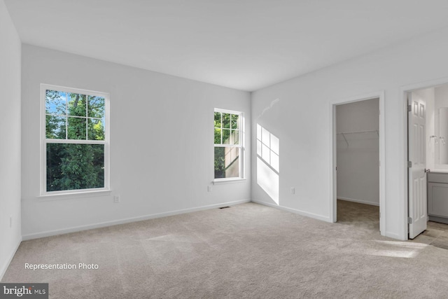 unfurnished bedroom featuring light carpet, a walk in closet, ensuite bath, and multiple windows