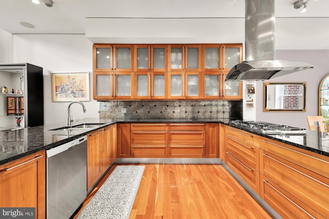 kitchen with sink, stainless steel appliances, dark stone countertops, light hardwood / wood-style floors, and island range hood