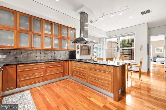 kitchen featuring decorative backsplash, dark stone countertops, island exhaust hood, and stainless steel gas cooktop