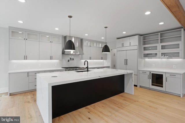 kitchen with paneled fridge, sink, wall chimney range hood, light hardwood / wood-style flooring, and an island with sink