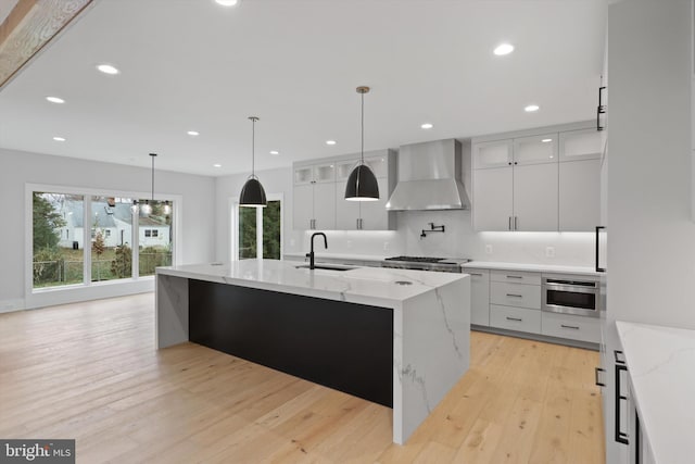 kitchen featuring a kitchen island with sink, white cabinets, wall chimney range hood, light wood-type flooring, and light stone counters