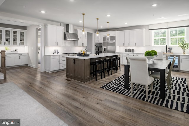 kitchen with white cabinetry, wall chimney range hood, stainless steel built in refrigerator, an island with sink, and pendant lighting