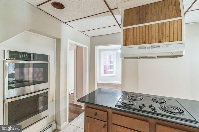 kitchen featuring a drop ceiling, black electric cooktop, baseboard heating, double oven, and light tile patterned floors