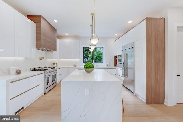 kitchen with wall chimney exhaust hood, pendant lighting, high quality appliances, white cabinets, and light wood-type flooring