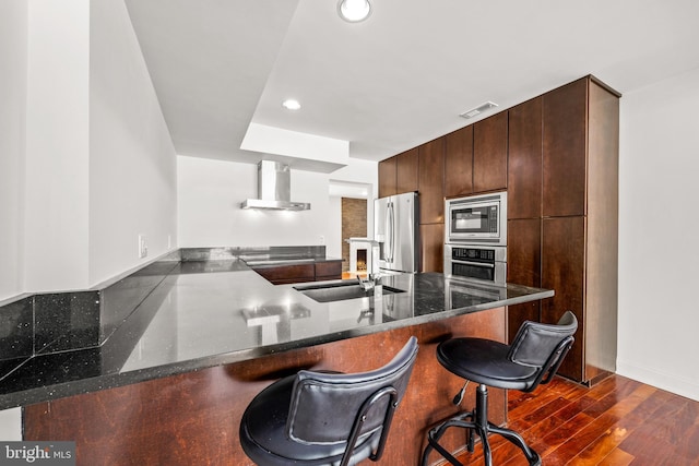 kitchen featuring wall chimney exhaust hood, dark hardwood / wood-style floors, kitchen peninsula, a breakfast bar, and appliances with stainless steel finishes