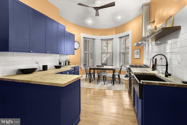 kitchen featuring wood counters, decorative backsplash, wall chimney exhaust hood, blue cabinetry, and light hardwood / wood-style floors