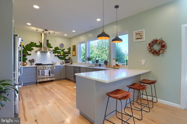 kitchen with light wood finished floors, appliances with stainless steel finishes, a peninsula, gray cabinets, and wall chimney range hood