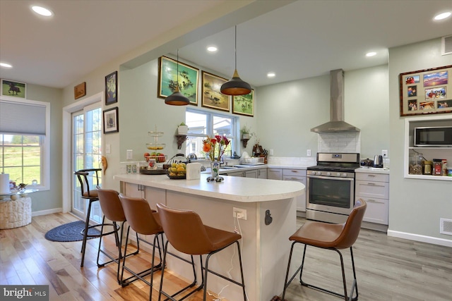 kitchen featuring a peninsula, stainless steel appliances, light countertops, a healthy amount of sunlight, and wall chimney range hood