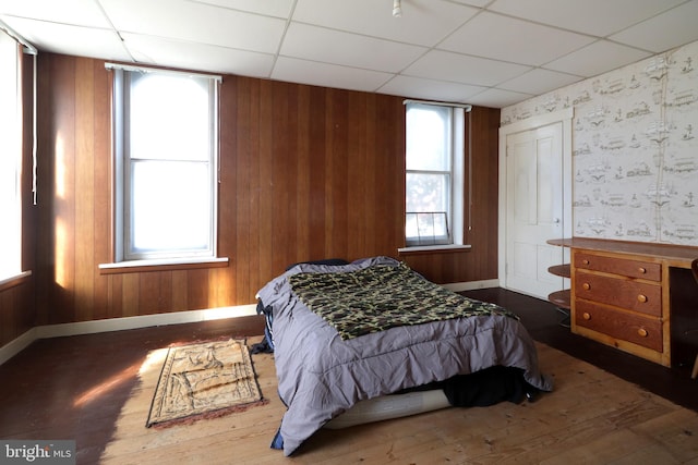 bedroom featuring dark hardwood / wood-style flooring, a drop ceiling, multiple windows, and wood walls