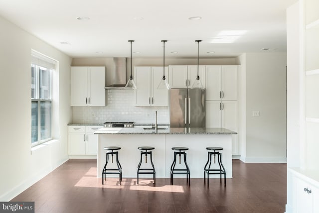 kitchen featuring a breakfast bar, a kitchen island with sink, a sink, freestanding refrigerator, and wall chimney range hood