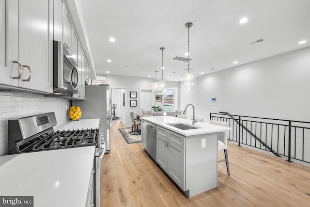 kitchen featuring sink, hanging light fixtures, stainless steel appliances, a kitchen island with sink, and light wood-type flooring