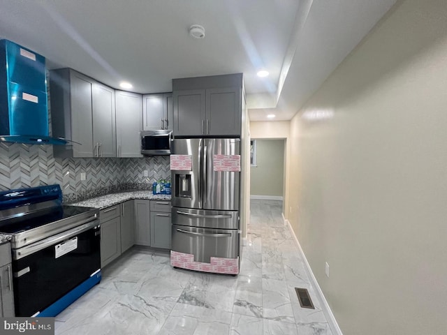 kitchen with wall chimney exhaust hood, gray cabinets, decorative backsplash, light stone countertops, and appliances with stainless steel finishes