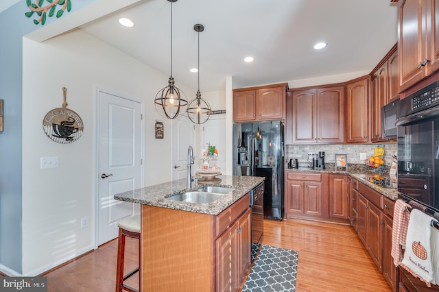 kitchen with sink, an island with sink, dark stone counters, light hardwood / wood-style floors, and black appliances