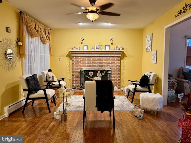 living area featuring hardwood / wood-style flooring, ceiling fan, a baseboard heating unit, and a brick fireplace