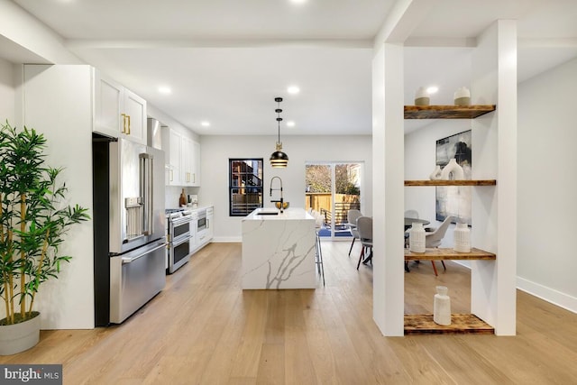 kitchen with light wood-type flooring, stainless steel appliances, a kitchen island with sink, white cabinets, and hanging light fixtures