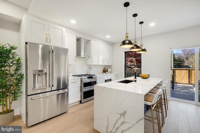 kitchen with a kitchen island with sink, white cabinets, wall chimney range hood, light stone countertops, and premium appliances