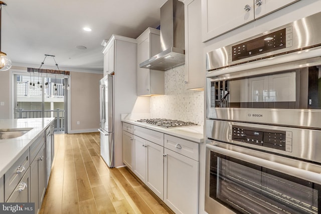 kitchen featuring wall chimney exhaust hood, ornamental molding, decorative light fixtures, appliances with stainless steel finishes, and light wood-type flooring