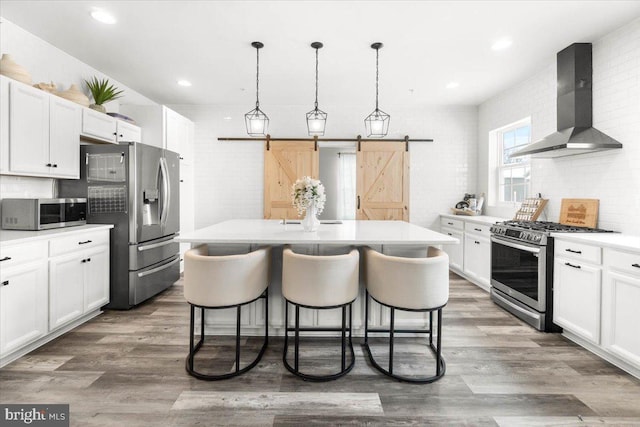 kitchen with appliances with stainless steel finishes, hanging light fixtures, wall chimney range hood, white cabinets, and a center island with sink