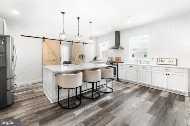 kitchen with wall chimney exhaust hood, decorative light fixtures, white cabinets, a center island with sink, and stainless steel appliances