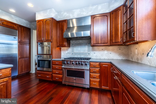 kitchen with decorative backsplash, premium appliances, dark wood-type flooring, and wall chimney exhaust hood