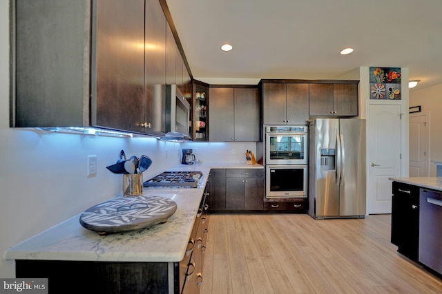 kitchen with dark brown cabinetry, stainless steel appliances, tasteful backsplash, light stone counters, and light hardwood / wood-style flooring