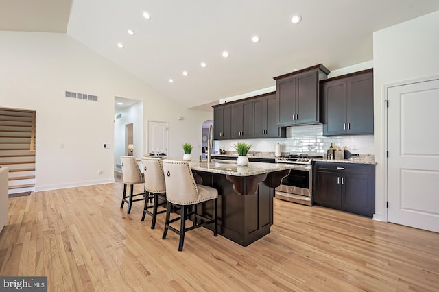 kitchen featuring backsplash, light stone counters, stainless steel gas range oven, a center island with sink, and light hardwood / wood-style floors