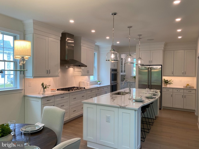 kitchen featuring sink, wall chimney range hood, an island with sink, appliances with stainless steel finishes, and light wood-type flooring