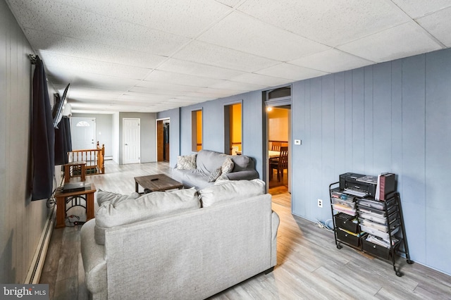 living room featuring light wood-type flooring, a baseboard radiator, a drop ceiling, and wood walls