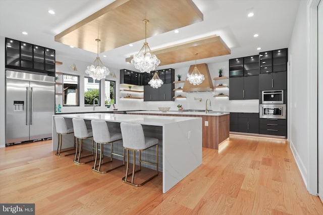 kitchen with open shelves, a tray ceiling, built in appliances, dark cabinets, and a center island