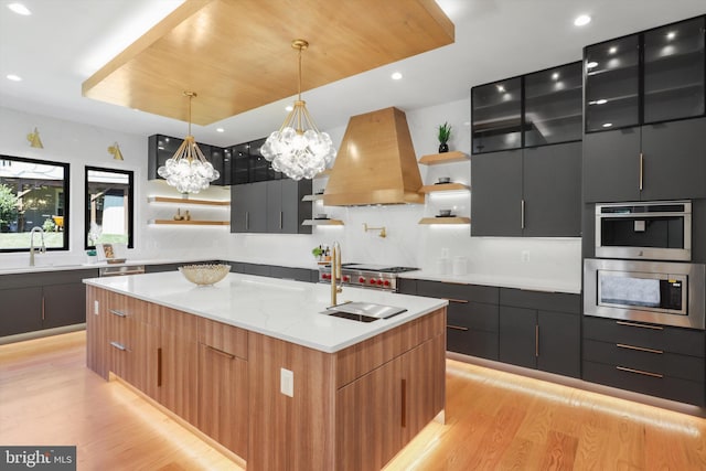 kitchen with premium range hood, light hardwood / wood-style flooring, hanging light fixtures, and a kitchen island with sink