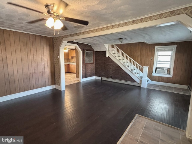 unfurnished living room with ceiling fan, a baseboard heating unit, wood walls, hardwood / wood-style flooring, and ornamental molding