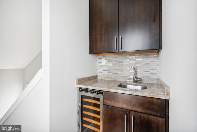 bar with decorative backsplash, light stone counters, dark brown cabinetry, sink, and wine cooler