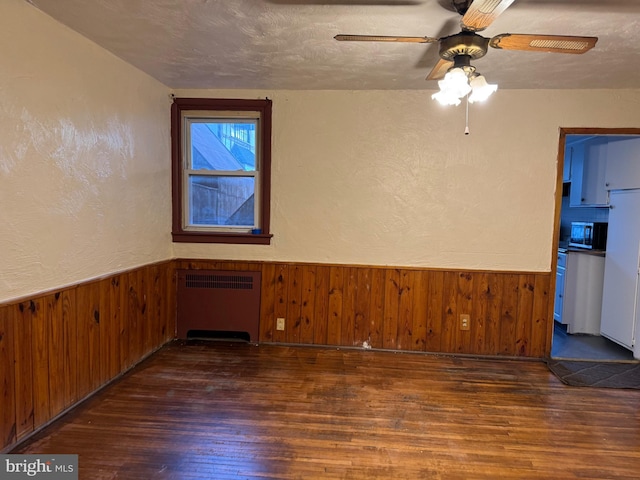 spare room featuring a textured ceiling, ceiling fan, wooden walls, dark wood-type flooring, and radiator heating unit