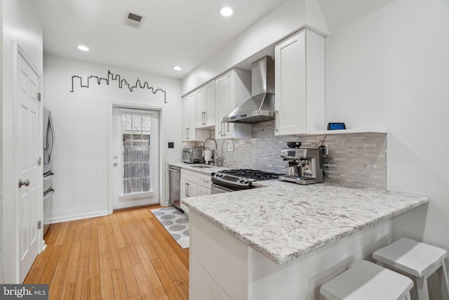 kitchen with white cabinets, wall chimney range hood, sink, light wood-type flooring, and appliances with stainless steel finishes