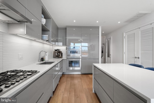 kitchen featuring exhaust hood, sink, gray cabinets, light wood-type flooring, and appliances with stainless steel finishes