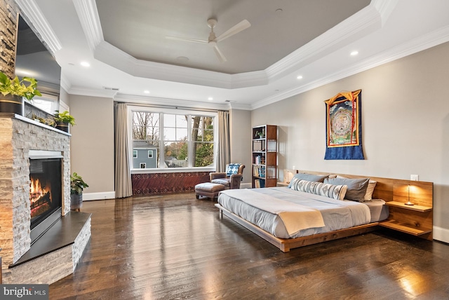 bedroom featuring a stone fireplace, ceiling fan, a raised ceiling, dark wood-type flooring, and ornamental molding