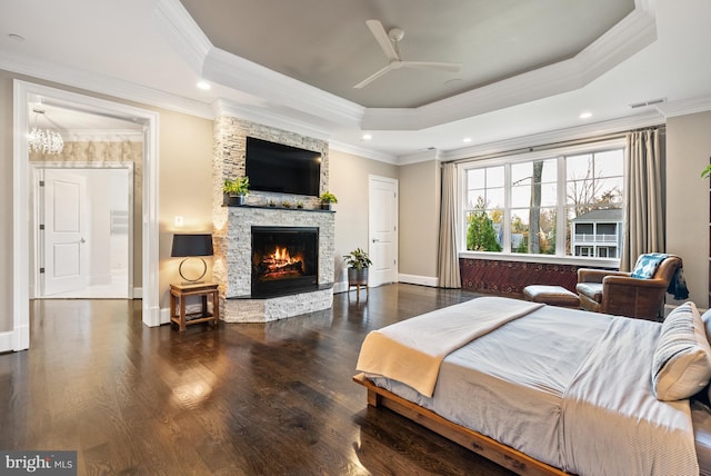 bedroom featuring a stone fireplace, ceiling fan, a tray ceiling, dark wood-type flooring, and ornamental molding