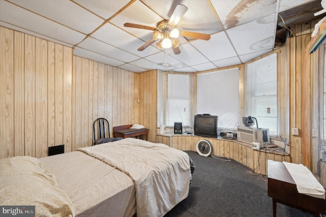 bedroom featuring a drop ceiling, wood walls, cooling unit, carpet flooring, and ceiling fan