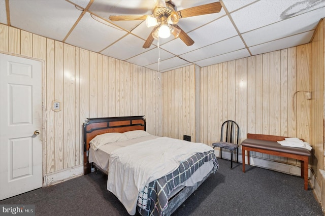 bedroom featuring ceiling fan, a drop ceiling, wood walls, and dark colored carpet