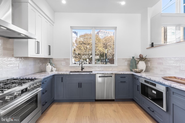 kitchen with sink, stainless steel appliances, wall chimney range hood, white cabinets, and light wood-type flooring