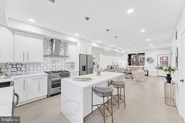 kitchen featuring pendant lighting, wall chimney exhaust hood, light stone countertops, appliances with stainless steel finishes, and white cabinetry