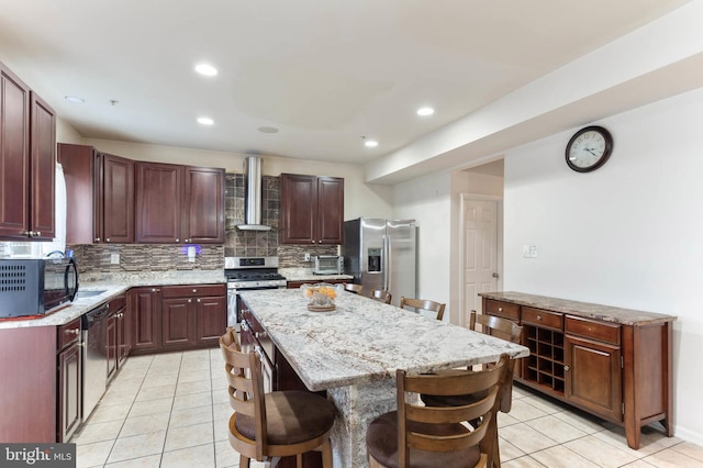 kitchen featuring wall chimney exhaust hood, stainless steel appliances, a kitchen breakfast bar, backsplash, and a kitchen island