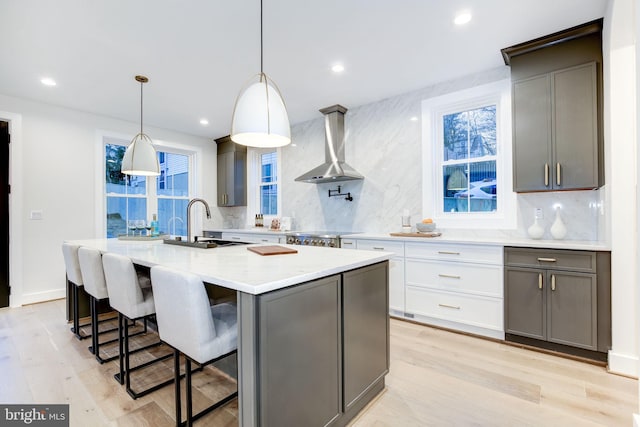 kitchen with gray cabinetry, white cabinetry, decorative light fixtures, an island with sink, and wall chimney range hood